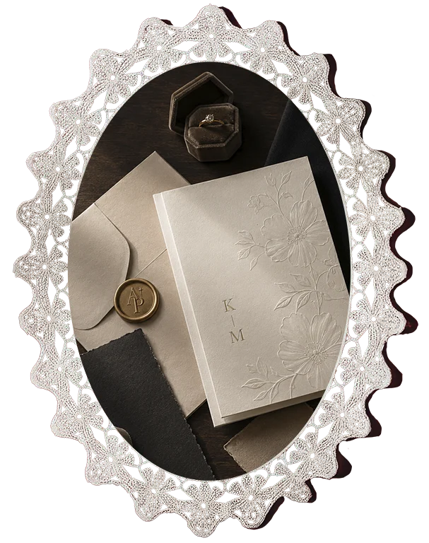 Oval lace-framed wedding stationery flat lay with textured paper, envelope, wax seal, and ring box
